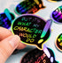 Hand holding a holographic speech bubble sticker that reads “But it’s what my character would do” in colorful text.