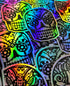 Pile of vibrant holographic skull stickers with black tribal designs and crown details, glowing in rainbow colors under light.